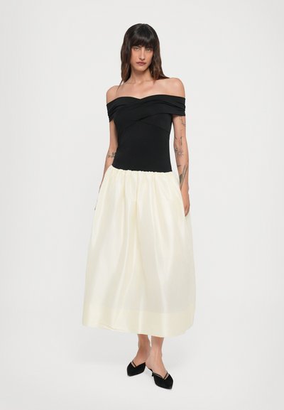 Sort off-shoulder top med rynke detaljer, parret med en flydende hvid midi-nederdel lavet af let stof, præsenteret med sorte flade sko.
