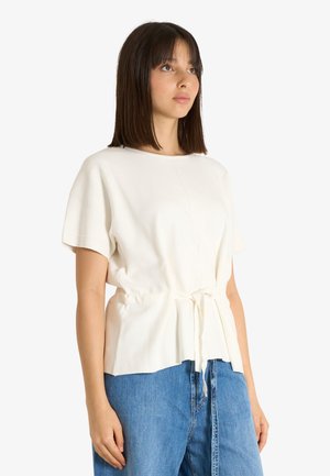 Donna con capelli castani lisci, che indossa una maglietta bianca a maniche corte con fiocco in vita e jeans blu, guardando leggermente verso l’alto di lato.