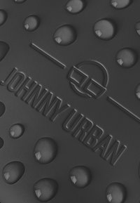 Svart texturerad yta med upphöjda droppar och den präglade texten "JOHNNY URBAN", med ett cirkulärt designmotiv.