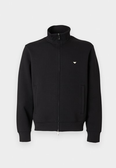 Juodas megztinis su užtrauktuku ir aukštu apykakliu, su dviem priekinėmis kišenėmis ir mažu auksiniu emblematu kairėje krūtinės pusėje. Minkštos audinio tekstūros.