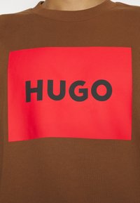 T-shirt en coton marron avec un grand motif rectangulaire rouge portant le texte "HUGO" en lettres noires en gras au centre.