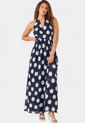 Frau mit lockigem Haar, die ein marineblaues Halter-Maxikleid mit großen weißen Polka-Dots und weiße, Riemchen-absatzsandalen trägt.