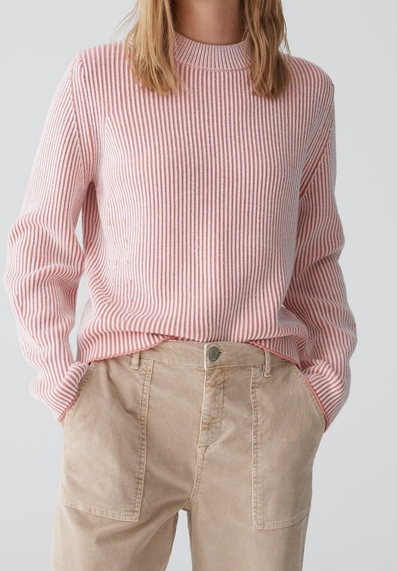 Pull côtelé à rayures rose clair et blanc avec un col haut, porté avec un pantalon en velours côtelé beige, équipé de poches latérales et d'un détail bouton.