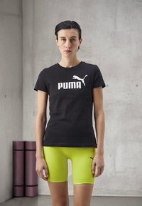 Juoda medvilninė marškinėliai su balta "PUMA" logotipu, derinami su kūną prigludusiais neoninės geltonos spalvos šortais. Paprastas, atletinis dizainas su lygiu paviršiumi.
