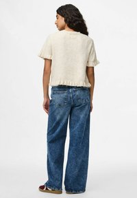 Bežový svetr s krátkým rukávem a volánkovými okraji, kombinovaný s volnými modrými džíny z denimu. Džíny mají klasický design s pěti kapsami.