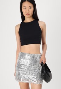 Top corto senza maniche di colore nero e una mini gonna argentata con paillettes e dettagli in frange. La modella tiene una borsa a tracolla nera in un tessuto morbido.