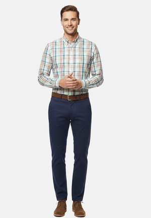 Hombre sonriente de pie con las manos entrelazadas, vestido con una camisa de cuadros abotonada, pantalones azul marino, cinturón marrón y zapatos de ante marrones, sobre fondo blanco.