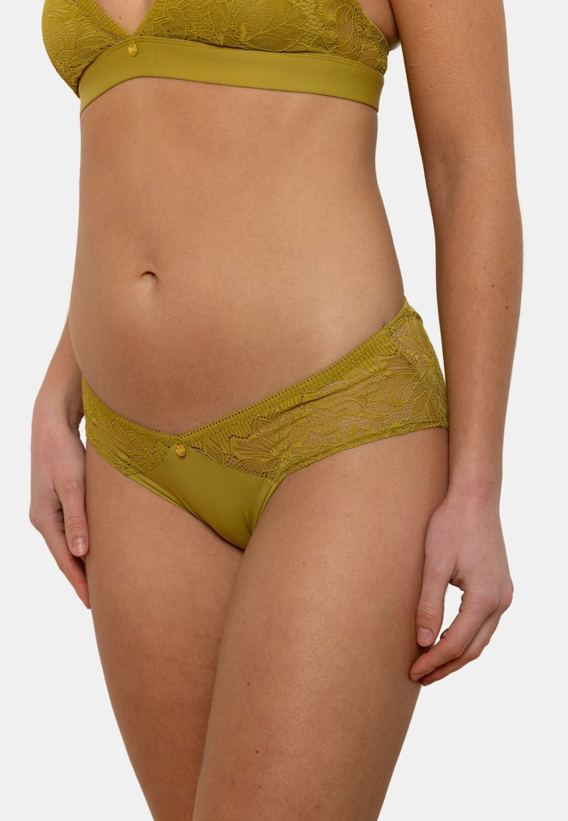 String vert olive avec des détails en dentelle sur les côtés, doté d'une taille lisse et d'un petit bouton décoratif à l'avant.