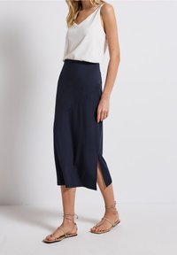 Tmavě modrá midi sukně s přiléhavým střihem, bočními rozparky a hladkou texturou. Kombinováno s bílým tílkem bez rukávů a plochými sandály.