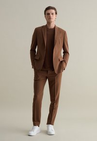Completo marrone su misura con camicia abbinata, tessuto leggero, pantaloni slim-fit e sneakers bianche. Presenta una tasca sul petto e linee nette.