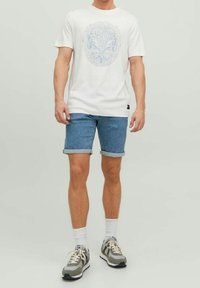Camiseta blanca de manga corta con estampado gráfico azul, combinada con pantalones cortos de mezclilla azul claro con puños enrollados y zapatillas grises con acentos beiges.
