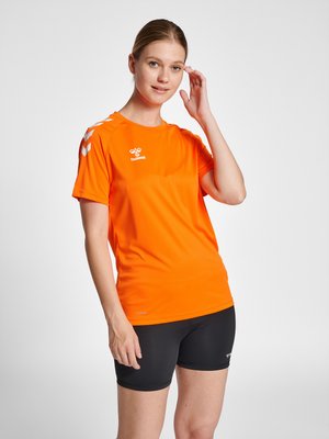 Vrouw in oranje sportshirt met korte mouwen en zwarte korte broek, haar haar rechtzettend, staand tegen een effen lichte achtergrond.