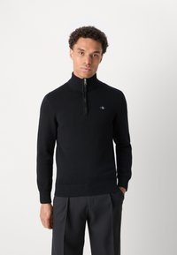 Mann steht mit einer Hand in der Tasche, trägt schwarzen Halb-Zip-Pullover und dunkle Hose vor einfachem weißem Hintergrund.