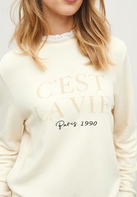 Beżowa bluza z napisem "C'est la vie" w pogrubionym jasnobrązowym kolorze oraz "Paris 1990" w czarnej kursywie, z teksturowanym wykończeniem materiału.