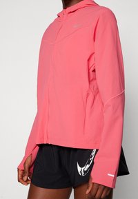 Veste Nike rose clair avec capuche, fermeture éclair, manches longues avec passe-pouces et poches latérales, fabriquée en matériau lisse et léger.