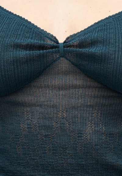 Σκούρο τιρκουάζ πλεκτό τοπ με V-λαιμόκοψη, υφή υφάσματος με διακριτική ραβδώσεις και κεντρική λεπτομέρεια κόμβου μπροστά. Μαλακό και ελαφρώς διάφανο.