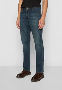 Dark blue denim jeans with a straight leg design, featuring five pockets and contrast stitching, worn with brown dress shoes.