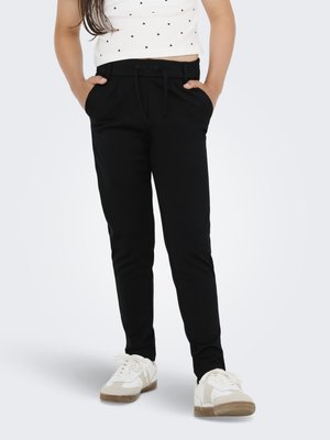 Kind trägt schwarze Hosen mit Kordelzug, weiße Sneaker und ein weißes Crop-Top mit kleinen schwarzen Herzmustern, Hände in den Taschen.