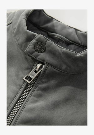 Veste matelassée grise avec un col montant, fermeture éclair métallique argentée et un accent en bouton rond au niveau du cou. Texture lisse, design élégant.