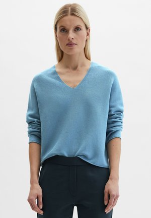 Blonde vrouw die een lichtblauwe V-hals trui draagt met opgerolde mouwen en donkere broek, staand tegen een effen witte achtergrond.