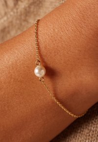 Gold chain bracelet featuring a single white pearl accent, set against tanned skin, showcasing a delicate and minimalistic design.