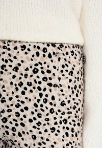 Schmale Hosen aus einem beigen Stoff mit einem schwarzen und braunen Leopardenmuster. Sie verfügen über einen sichtbaren Metallknopf und eine Nahtdetails.