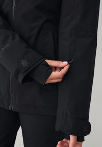 Zwarte jas gemaakt van glad, waterdicht materiaal; voorzien van een ritszak op de mouw en een klittenbandsluiting bij de manchetten. Subtiele branding op de arm.