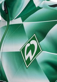 Grön och vit mönstrad sportjacka med en distinkt logotyp och en dragkedja vid kragen. Sllät, lättviktig tyg med geometrisk design.