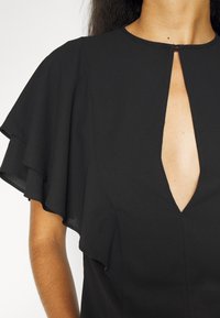 Femme portant un top noir avec des manches courtes superposées et flottantes, et un décolleté en trou de serrure à l'avant maintenu par un petit crochet.