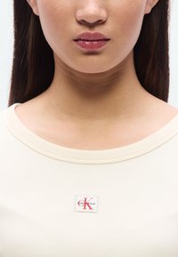 Ανοιχτό μπεζ ριμπ βαμβακερό τοπ με στρογγυλή λαιμόκοψη και τετράγωνο λογότυπο Calvin Klein με κόκκινες και μαύρες λεπτομέρειες.