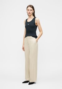 Γυναίκα που φοράει μαύρο αμάνικο τοπ, μπεζ παντελόνι φαρδιάς γραμμής και μαύρες μυτερές μπαλαρίνες, στέκεται με το ένα χέρι στην τσέπη.