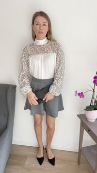 Donna che indossa una blusa di pizzo bianco, una gonna a scacchi, scarpe nere a punta, mentre tiene una pochette nera, in piedi all'interno accanto a una poltrona grigia e a un tavolo con un'orchidea.