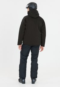 Schwarze Skijacke mit Kapuze, getragen über dunklen Hosen. Merkmale sind eine glatte Textur und ein tailliertes Design. Ein schwarzer Helm vervollständigt das Outfit.