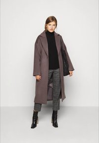 Nuori nainen, jolla on pitkä mauve-takki, musta poolopaita, harmaan ruudulliset housut ja mustat nilkkurit, sekä hän kantaa mustaa laukkua olkapäällä.