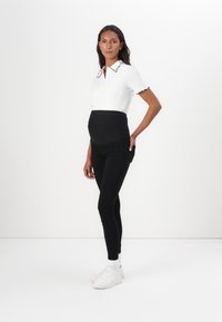 White polo shirt with black scalloped collar, paired with black maternity leggings and white sneakers. Model stands against a plain background.