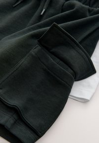 Pantalon de survêtement vert foncé à la texture douce, doté d'une taille élastique et de grandes poches latérales ; t-shirt blanc partiellement visible en dessous.