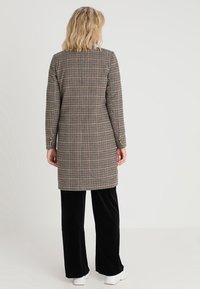 Cappotto lungo a quadri nei colori marrone, crema e nero. Presenta maniche lunghe, un taglio dritto e dettagli con bottoni vicino ai polsini. Abbinato a pantaloni neri.