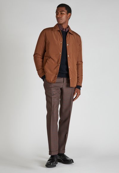 Man in een bruin jack, marineblauwe sweater, geruite bruine broek en zwarte schoenen die tegen een neutrale achtergrond staat, met zijn handen in zijn zakken.