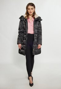 Geacă puffer neagră cu glugă căptușită cu blană sintetică, fermoar la față și buzunare laterale, purtată peste o bluză roz și pantaloni negri, cu o textură fină.
