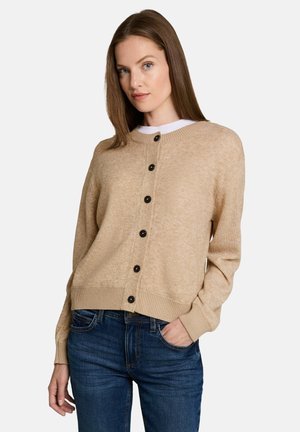 Woman wearing a beige buttoned cardigan over a white shirt and blue jeans, standing with one hand in her pocket against a plain background.