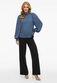 Kék pulóver, textúrált anyaggal, magas nyakkal és puffos ujjal, fekete, bő szabású nadrággal és pántos arany magassarkúval párosítva.