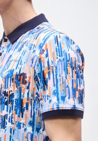 Άνδρας που φοράει κοντομάνικο πόλο πουκάμισο με κάθετες ρίγες σε μπλε, πορτοκαλί και λευκό χρώμα και λουλουδάτα σχέδια, καθώς και σκούρο γιακά και μανσέτες στα μανίκια.