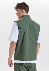 Zelená bezrukávová vesta vyrobená z lehkého materiálu má stojáček a nosí se přes bílou košili s krátkým rukávem. Minimalistický design.