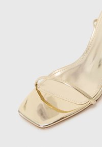 Sandale à talons dorée en métal avec un bout carré, présentant des fines sangles sur le pied et une texture lisse et brillante.