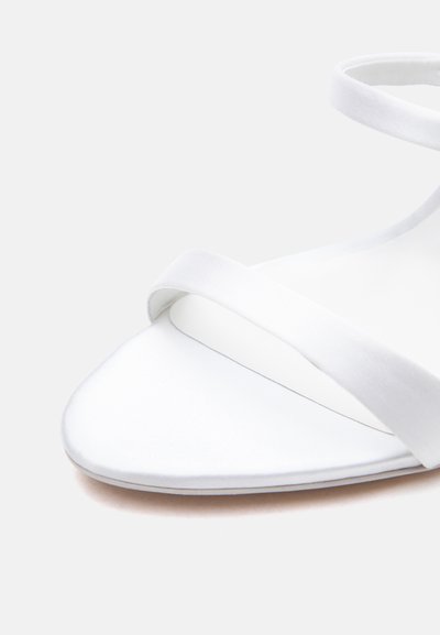 Λευκό πέδιλο ανοιχτού δακτύλου με λεπτά λουράκια που διασταυρώνονται πάνω στο πόδι και γύρω από τον αστράγαλο σε ουδέτερο φόντο.