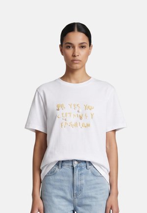 Woman with dark hair tied back wears white T-shirt with yellow text "OH YES YOU CERTAINLY FASHION" and light blue jeans against white background.