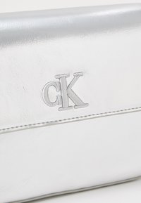Bílá kožená kabelka na clutch s hladkým povrchem, s vyšitým stříbrným logem "cK" a jemným prošitím podél okraje.