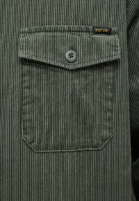 Grünes Cordhemd mit Brusttasche, Knopfverschluss und vertikaler Rippstruktur. Etikett nahe der Schulter eingenäht.