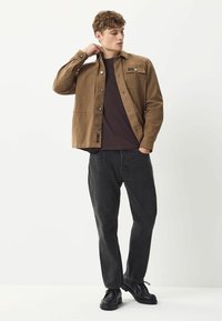 Giacca di cotone marrone con bottoni a pressione, una tasca sul petto, indossata sopra una camicia scura. Abbinata a jeans neri e scarpe nere.
