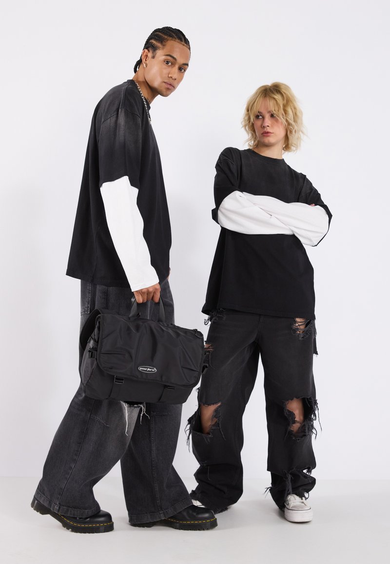 Bolso mensajero negro con correa ajustable, que presenta una textura suave y un diseño de logo, llevado con jeans negros de gran tamaño y rasgados, y mangas de contraste.